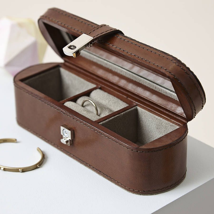 The image shows an open, brown leather jewelry box placed on a white surface. The box has a curved, vintage design with a metal lock and stitching details. 