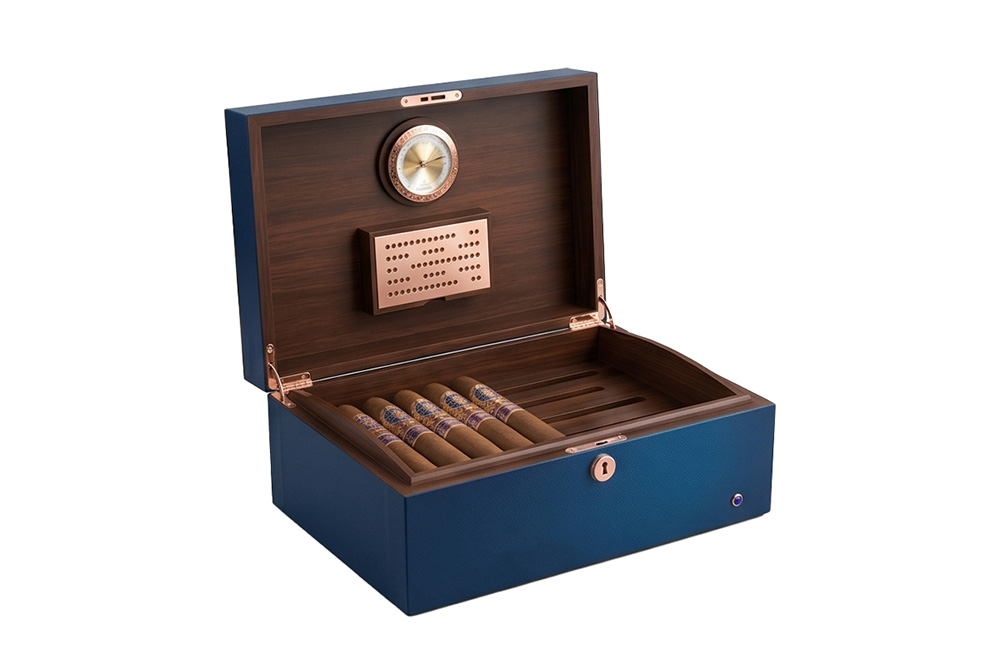 Un humidificateur à cigares ouvert, luxueux, en cuir bleu et bois, vu de biais, mettant en valeur la doublure en cèdre espagnol, l’hygromètre et l’humidificateur du couvercle, ainsi que des cigares disposés dans le plateau inférieur.