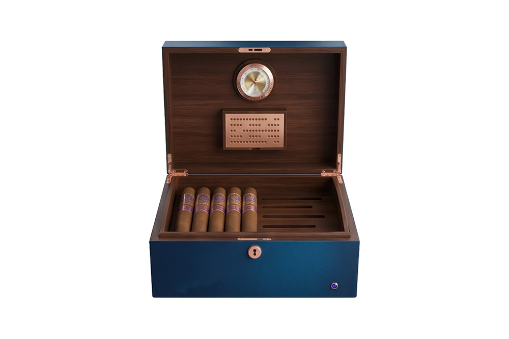 Humidor à cigares haut de gamme en cuir bleu et bois de cèdre espagnol, vue de face, mettant en évidence l’hygromètre doré intégré, l’humidificateur métallique et des cigares soigneusement rangés dans un plateau, isolé sur fond blanc